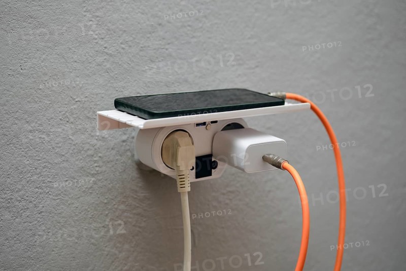 A smartphone rests on a wall-mounted charging station with an electrical outlet below