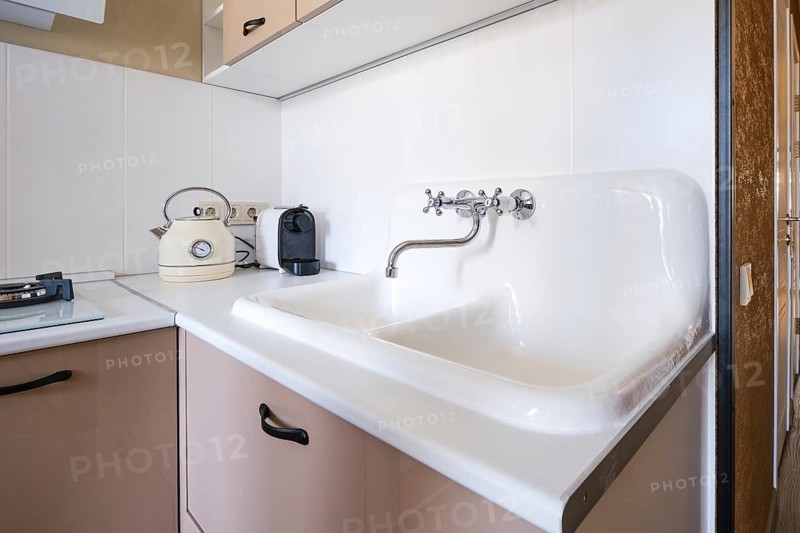 A clean kitchen space features a double sink with a vintage kettle beside a sleek coffee maker. Sunlight brightens the area, highlighting the minimalist design and functional layout