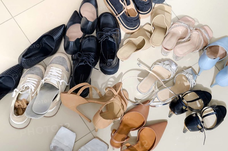 A collection of diverse footwear types is arranged on a light-colored tiled floor. Styles include sneakers, sandals, heels, and flats, showcasing different colors and designs in a playful manner