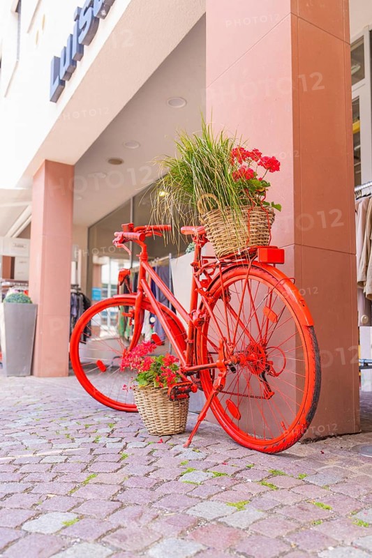 A red bicycle with flower baskets stands decoratively on a pavement in front of shops, small town of Perle Calw, Black Forest, Germany