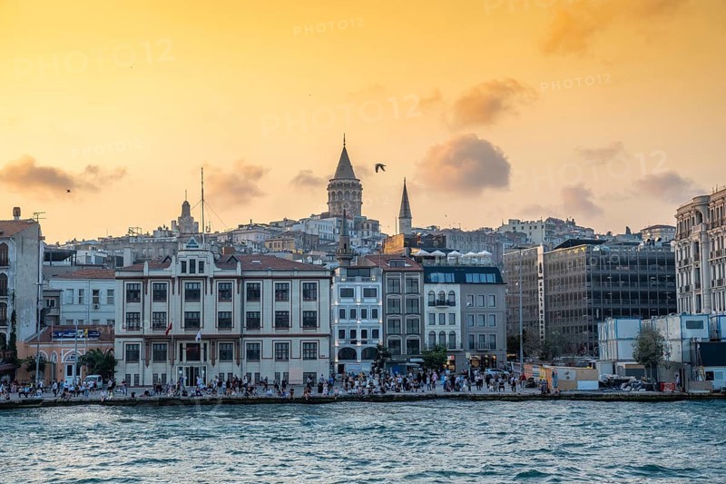 Tourists enjoying sunset view of galata tower and cityscape of istanbul, turkey, from the bosphorus strait with golden light illuminating buildings and cloudy sky