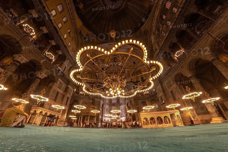 Tourists gather beneath the magnificent chandeliers within the hagia sophia, a historical landmark in istanbul, turkey, experiencing its architectural grandeur