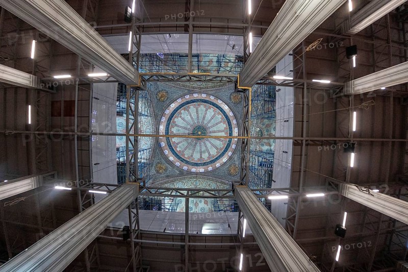 Wide angle view capturing ongoing restoration efforts within a mosque, highlighting scaffolding encasing a beautifully decorated dome, showcasing intricate details and the scale of the project