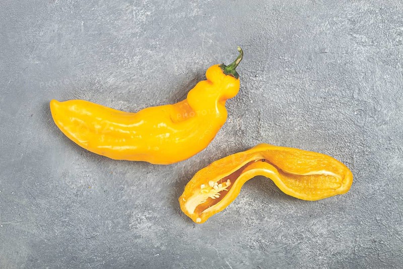 Two unusual yellow peppers are displayed on a textured gray surface. One pepper is whole while the other is cut in half, revealing its inner seeds and bright flesh, highlighting their unique shapes