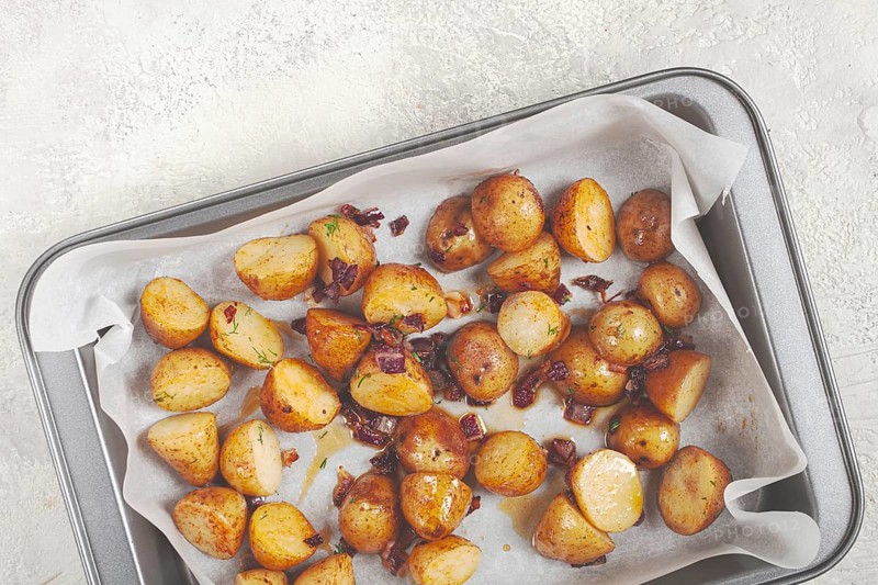 Golden-brown roasted potatoes are arranged on a baking tray lined with parchment paper. They are seasoned with herbs and garnished with crispy bacon bits, ready to be enjoyed