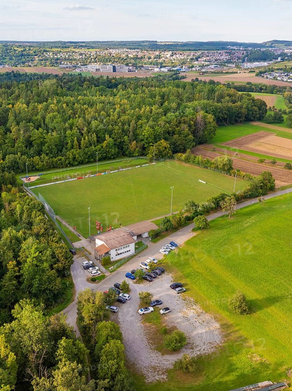 Aerial view of a sports field in the middle of wooded and rural surroundings with parked cars and a view of a village, VFL Stammheim, Calw, Black Forest, Germany