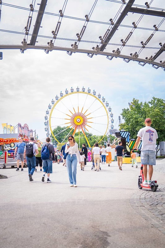 People in amusement park with e-scooter and big Ferris wheel in the background, Olympiapark, Munich, Germany