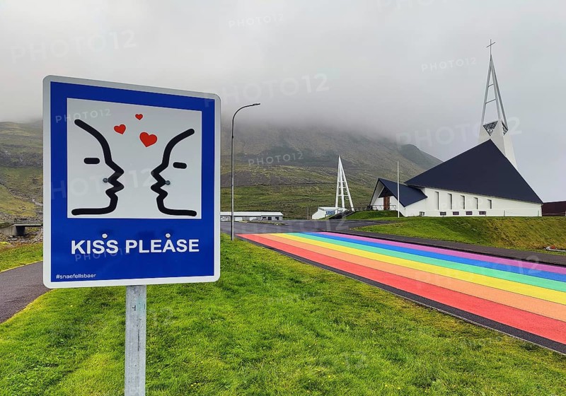 Sign with the invitation to kiss with a street painted in the colours of the rainbow and the church, Ólafsvík, Snæfellsnes, Iceland
