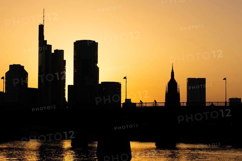 Two cyclists pass the rafter bridge over the Main in the evening while the sun sets behind the Frankfurt banking skyline, Frankfurt am Main, Hesse, Germany
