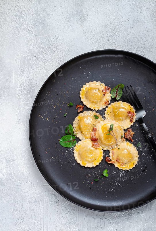 Fresh ravioli, with butter, spices and herbs, fried bacon, on a black plate, top view, dark background, no people
