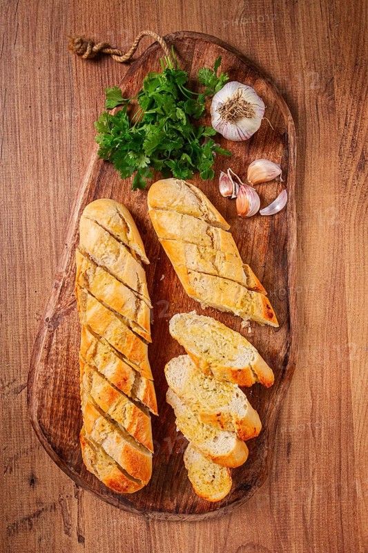 Garlic bread, baguette with garlic butter and herbs, on a wooden board, with herbs and garlic cloves