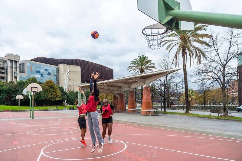 Diverse group of friends enjoying a casual game of street basketball on a red court, actively jumping and attempting to score under a cloudy sky in a modern urban environment