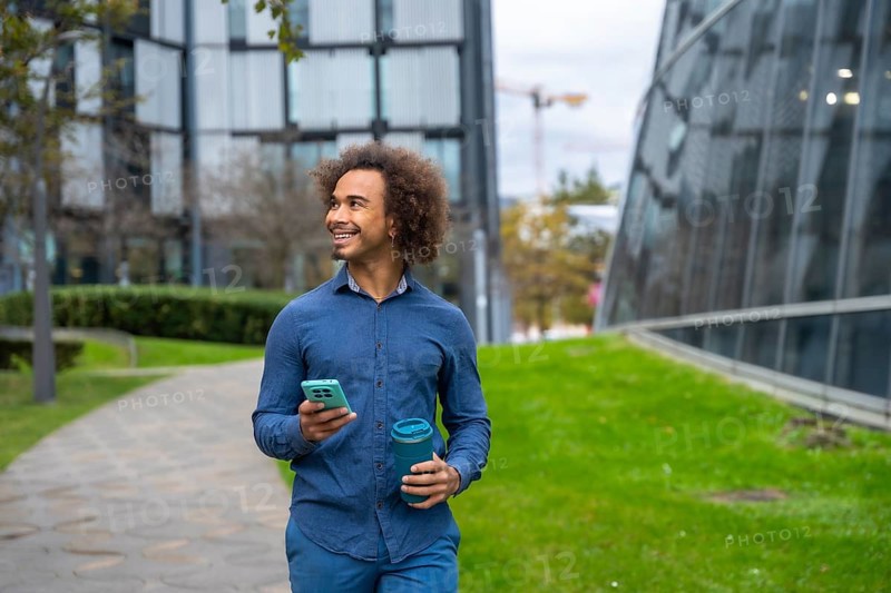 Smiling young Mixed-race man with curly afro hair walking on a path in a modern city park, holding a smartphone and a portable reusable cup, looking to the side