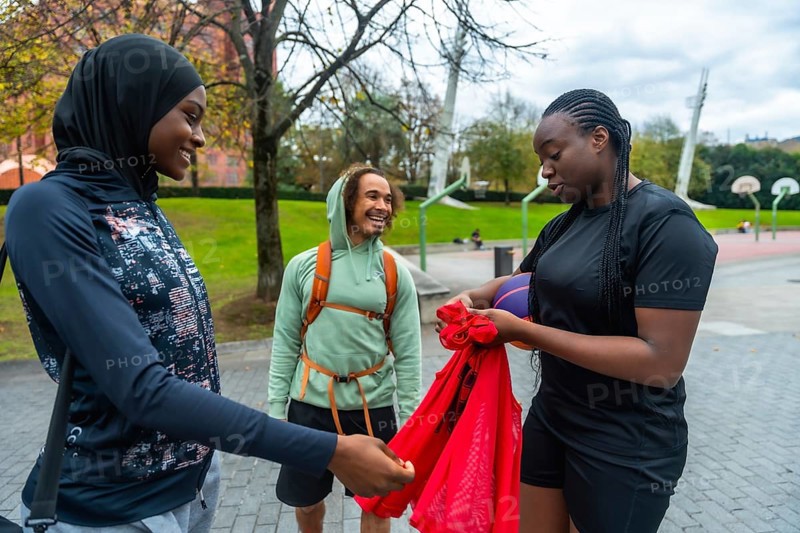 Three diverse friends, including two women wearing sportswear, are cheerfully gathering their gear on a basketball court, representing community, modern lifestyle, and friendship