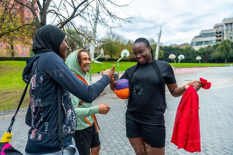 Three diverse friendswoman in hijab, man with curly hair, and woman holding a colorful basketballcheerfully high five on an outdoor urban court, celebrating sport and friendship