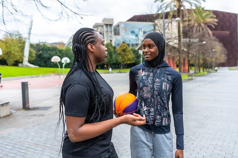 Diverse young women, one wearing a hijab and another with braids, talking and bonding while holding a basketball outdoors in an urban basketball court setting