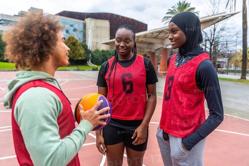 Young diverse athletes wearing sports bibs, holding a basketball, and talking on an urban court, reflecting teamwork, inclusion, and a healthy lifestyle