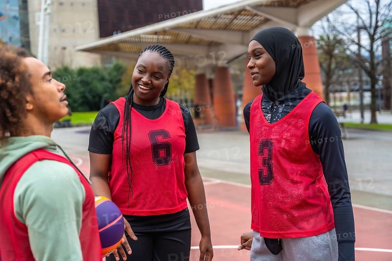 Group of diverse young athletes, including men and women, wearing red training bibs and a hijab, smiling and talking on an urban basketball court after playing a game