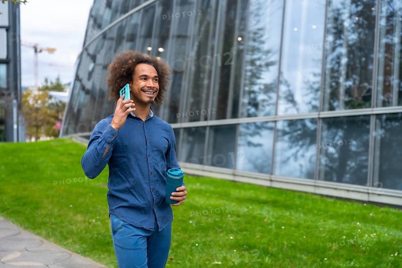 Smiling young Mixed-race man with curly hair walking confidently along a modern office building, holding a reusable cup and having a business conversation on his smartphone