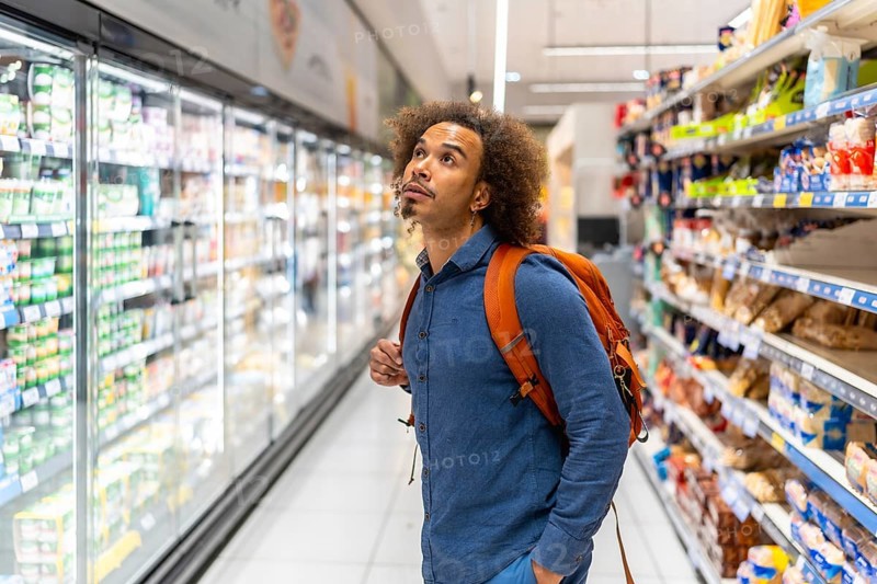 Young man with orange backpack studying refrigerated dairy products on supermarket shelves while walking the modern grocery aisle, contemplating choices during routine shopping trip