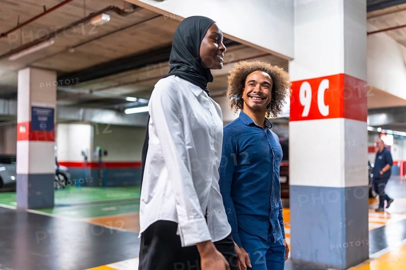 Diverse young couple walking happily and smiling together in a modern underground parking garage after parking their vehicle, representing urban transportation and connection