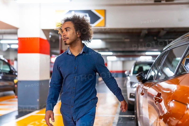Young man with afro hair walks through a brightly lit underground parking garage, glancing right as he searches for his car among rows of vehicles and concrete columns