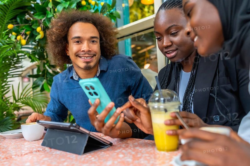 Young diverse friends in a cozy cafe sharing smartphone content, chatting and laughing over coffee and juice, relaxed together with a tablet and casual city vibe energy