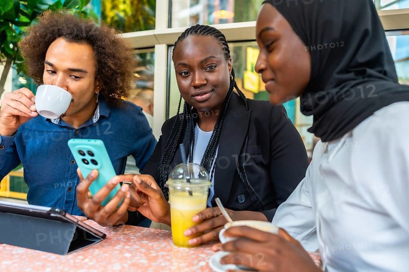 Group of multiethnic young adults relaxing together at a cafe, using a smartphone, discussing content and sharing social connection over beverages and a tablet on the table