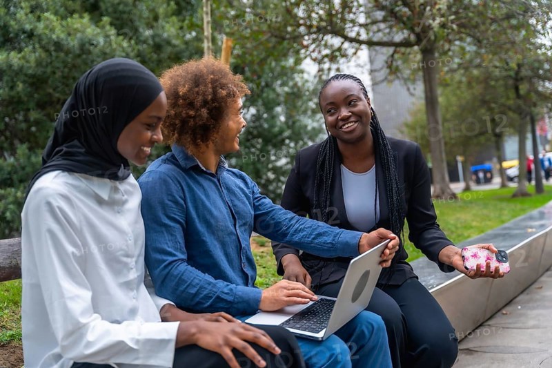 Diverse group of young adult students smiling and collaborating on a laptop in a city park, representing education, friendship, and digital learning outdoors