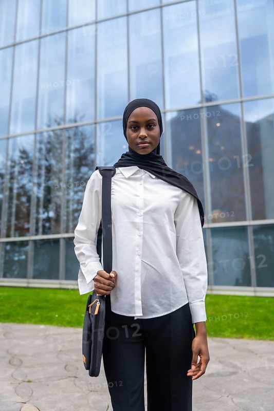 Young black muslim woman wearing a hijab and white shirt, holding a bag and standing confidently outside a contemporary glass building, representing education and successful futures