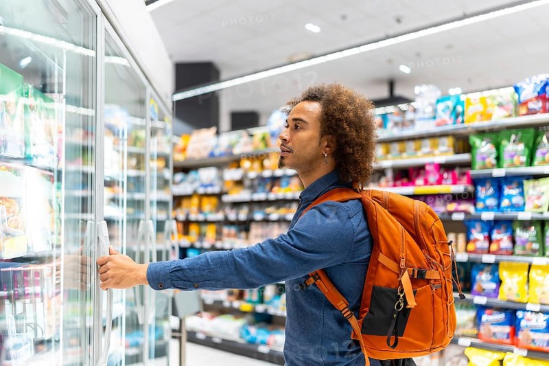 Young african american man with curly hair and a backpack opening a supermarket freezer, thoughtfully choosing frozen food while shopping in a modern grocery aisle