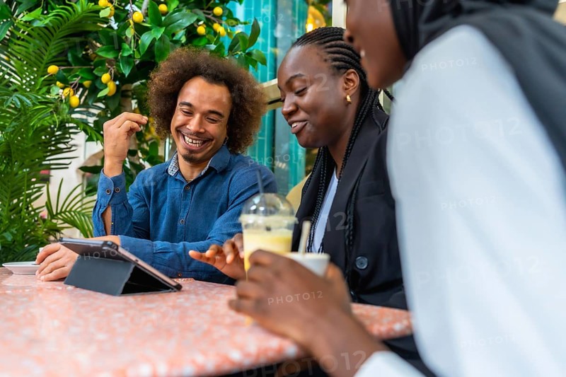 Cheerful diverse friends sitting at a cafe table, laughing and discussing content on a digital tablet while enjoying beverages, representing connection, leisure, and modern social interaction