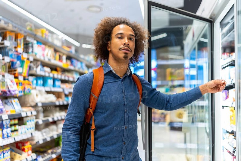 Young man with backpack selecting chilled items from a refrigerated display in a bright modern supermarket aisle, thoughtful consumer choosing fresh grab and go options