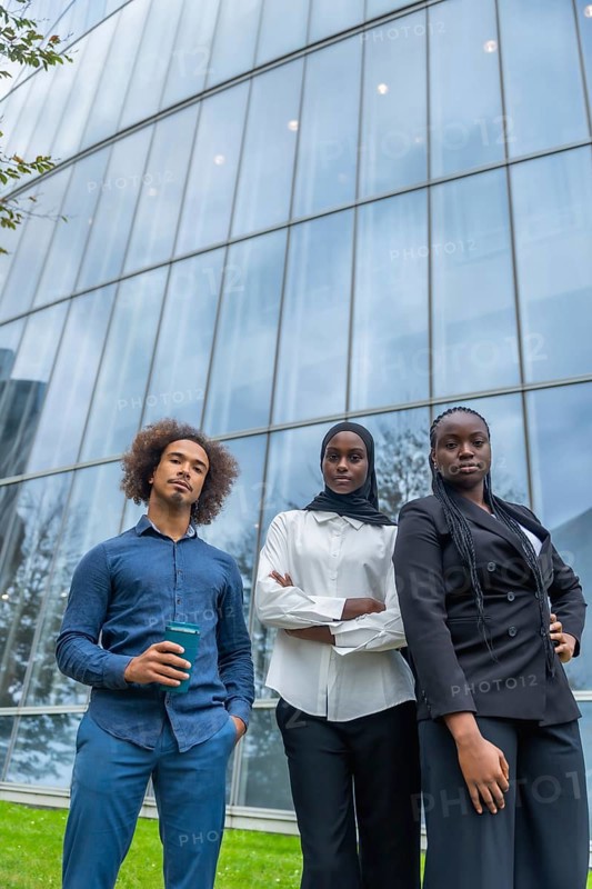Group of young diverse business professionals standing with confident serious expressions in front of a contemporary glass office building, representing corporate success and global teamwork