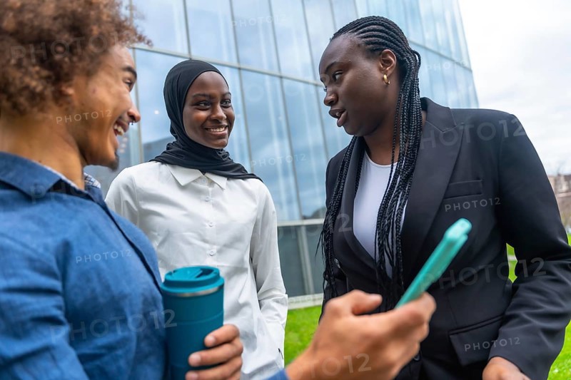 Group of multi ethnic colleagues engaging in a friendly conversation outside a modern office building, sharing ideas and using a mobile device for collaboration