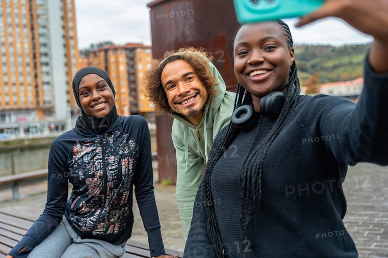 Three smiling diverse young adults enjoying friendship and making a selfie outdoors in an urban setting, wearing activewear and showing cheerful expressions