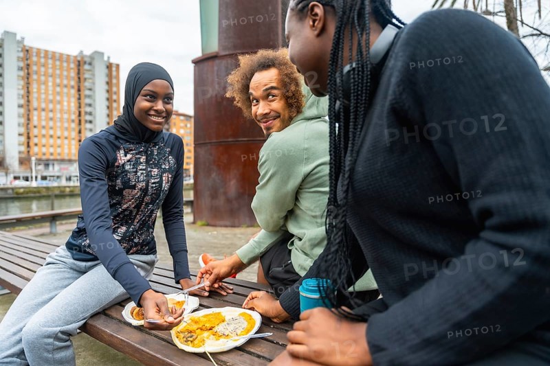 Diverse group of young adults sharing a casual outdoor meal on a city park bench, smiling, talking and relaxing together during an urban lunch or picnic break