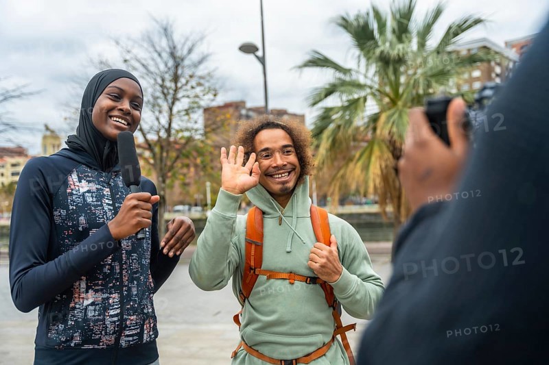 Smiling female journalist wearing a hijab holding a microphone, interviewing a happy man waving on the street while filming for a news report, capturing diverse content
