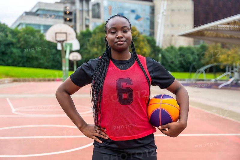 Confident young black woman athlete with braids wearing a red jersey, holding a basketball, and standing on an outdoor court with hands on hips, looking directly at the viewer