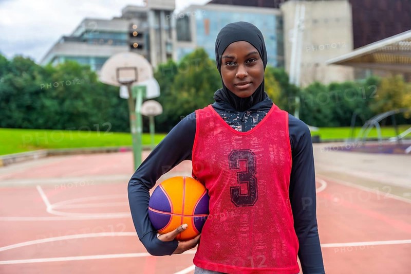 Young muslim woman wearing a hijab, long sleeve shirt, and basketball bib, holding a basketball while standing on an outdoor court, embodying confidence and athletic spirit