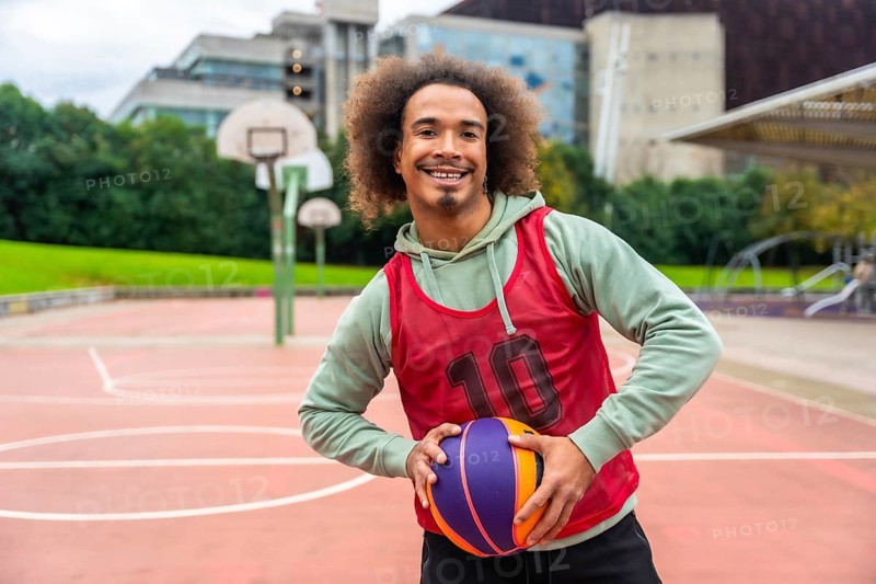 Young man with afro hairstyle smiling and holding a colorful basketball while standing on an outdoor urban basketball court, showing a positive and healthy lifestyle