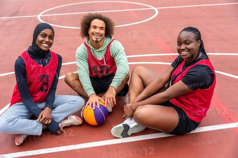 Diverse young friends wearing sport jerseys and a hijab, smiling while sitting together on an outdoor basketball court with a ball, representing teamwork and active youth