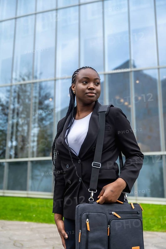 Confident black businesswoman standing outdoors, holding a briefcase, looking away while waiting, symbolizing professionalism, success, and ambition in a corporate urban environment