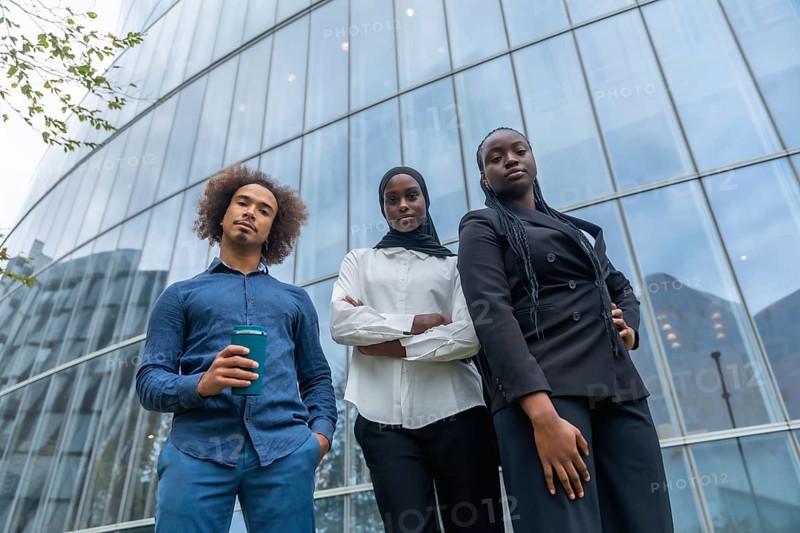 Diverse business team of young professionals standing together with confident expressions against a backdrop of a contemporary glass skyscraper, symbolizing ambition and success