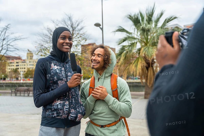 Female journalist interviewing a young, happy man with diverse background, holding a microphone while someone records a video outdoors in a city street