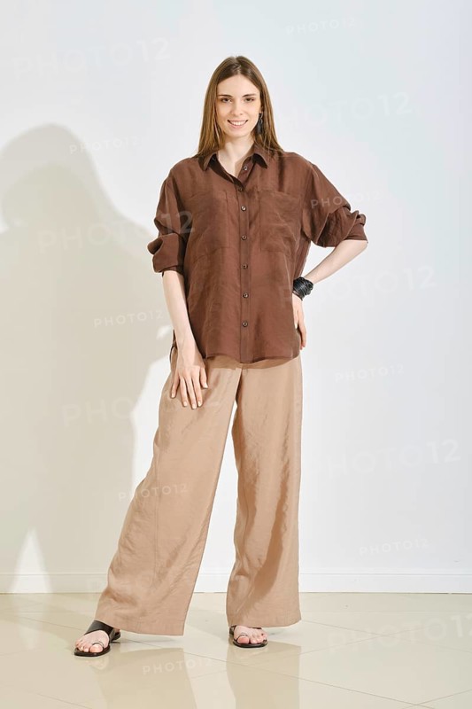 A woman in a trendy and practical outfit, featuring a brown shirt and loose cream trousers stands in bright room. The neutral colors and relaxed fit make it ideal for casual outings or everyday wear