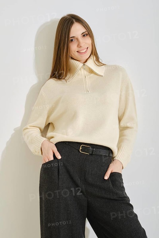 A woman stands by the bright wall with a warm smile, wearing a stylish cream sweater and loose dark trousers. Her outfit combines comfort and elegance, perfect for casual outings in a modern setting