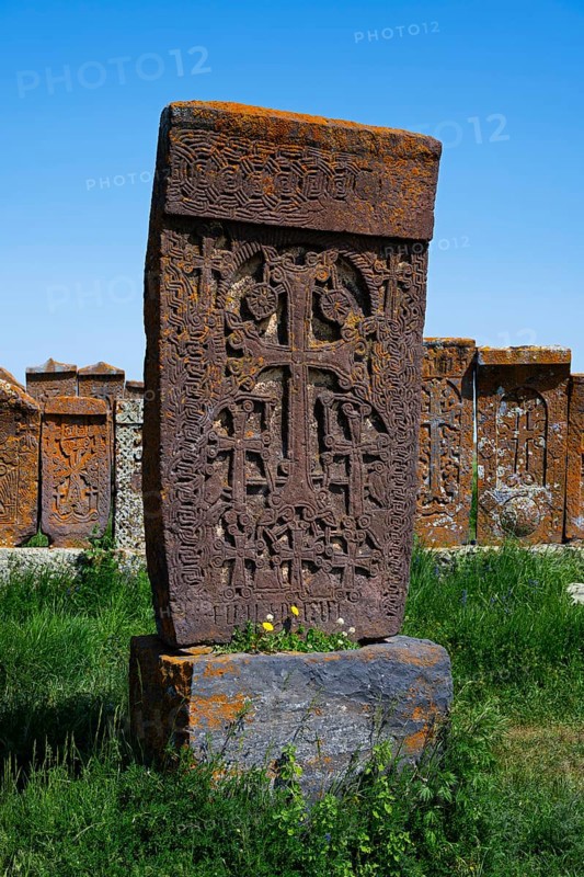 A detailed Armenian crossstone with engravings in front of several others, surrounded by grass under a clear sky, crossstones, khachkar, world's largest khachkar field, Noratus cemetery, Noradus, Gegharkunik province, Armenia