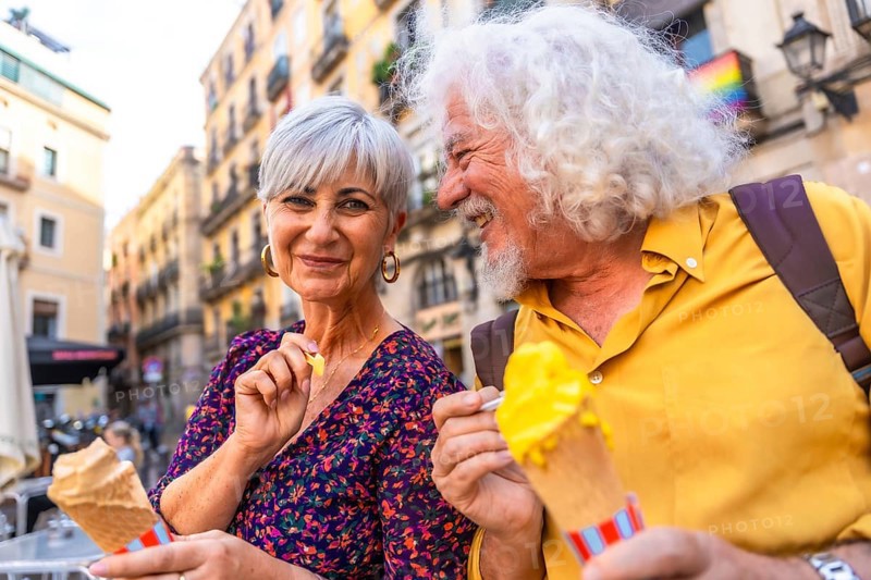 Happy mature couple smiling together while eating ice cream cones, experiencing leisure and love during a city break, with a pride flag representing inclusivity visible in the background