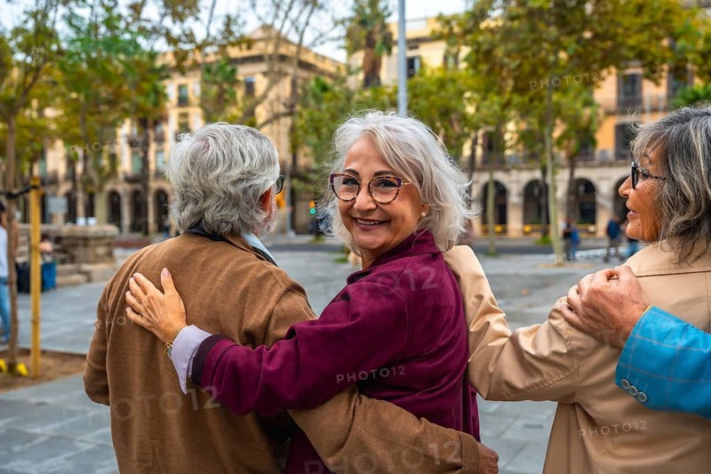 Group of three joyful senior friends walking together, hugging, and smiling at the camera in an urban public square, enjoying their shared bond and companionship